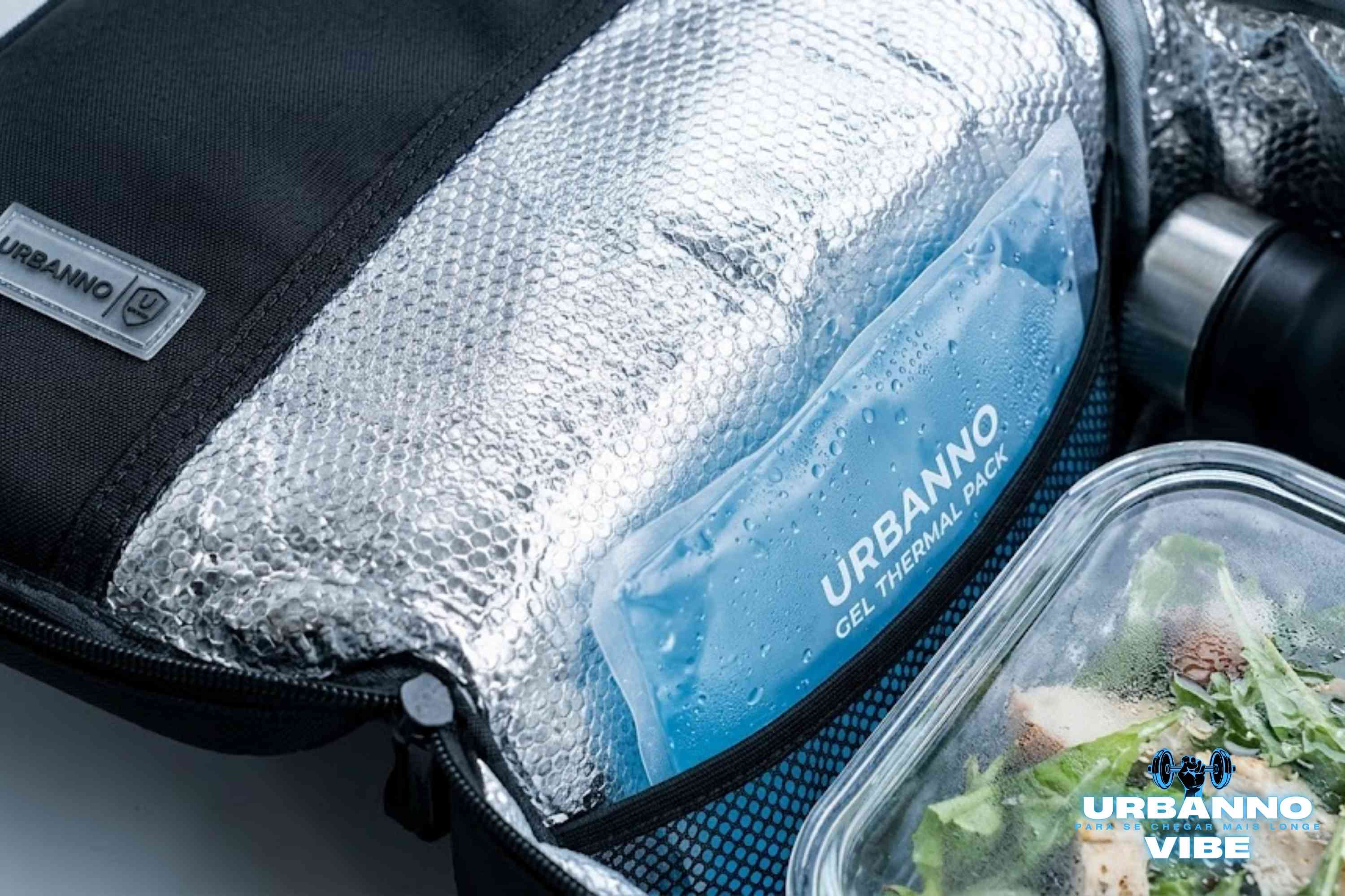 Visão interna em close (macro) de bolsa térmica Urbanno U-Armor mostrando revestimento de folha de alumínio, gel térmico azul suando de gelado e marmita de vidro com salada fresca.