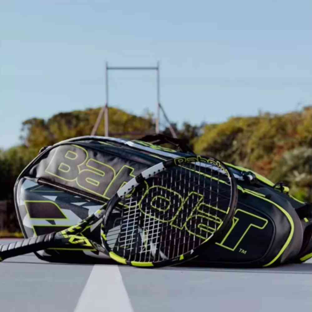 Raqueteira Babolat RH6 Pure Aero preta e amarela, vista de perto com uma raquete de tênis Babolat por cima, em uma quadra de tênis externa.