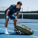 Jogador de tênis em quadra azul descansando, com a Raqueteira Babolat Pure Aero preta e amarela no chão. Foto de lifestyle em dia de sol.