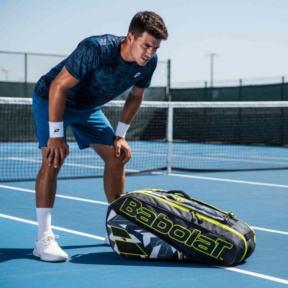 Jogador de tênis em quadra azul descansando, com a Raqueteira Babolat Pure Aero preta e amarela no chão. Foto de lifestyle em dia de sol.