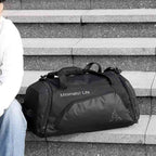 Bolsa de Academia Grande com Compartimento de Sapatos Preta Urbanno, com a inscrição "Minimalist Life", apoiada em degraus de concreto ao lado de uma pessoa sentada, ilustrando seu uso cotidiano.