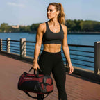 Mulher atlética usando top e calça preta, carregando a Bolsa de Academia Grande com Compartimento de Sapatos Vermelha Urbanno pela alça, em uma orla com água ao fundo, demonstrando seu uso em estilo de vida ativo.