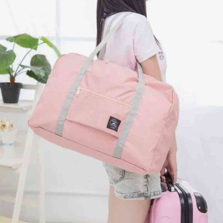 Bolsa de Academia e Viagens Dobrável Rosa Salmão Urbanno, sendo carregada no ombro por uma pessoa ao lado de uma mala rosa, demonstrando sua leveza e praticidade para viagens.