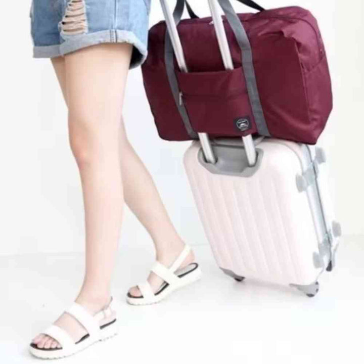 Bolsa de Academia e Viagens Dobrável Vinho Urbanno, acoplada à alça de uma mala de viagem clara, sendo transportada por uma pessoa, demonstrando sua praticidade para viagens.