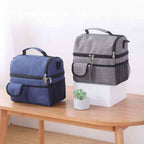 Duas Bolsas Térmicas Grandes para Marmita com Duplo Compartimento 10L Urbanno, uma azul e outra cinza, lado a lado em uma mesa de madeira com uma pequena planta, mostrando as opções de cores e design urbanno