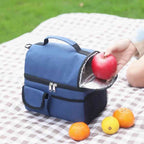  Bolsa Térmica Grande para Marmita com Duplo Compartimento 10L Urbanno azul, aberta em toalha de piquenique, com mão colocando uma maçã no compartimento superior, cercada por frutas cítricas, ideal para lazer ao ar livre urbanno