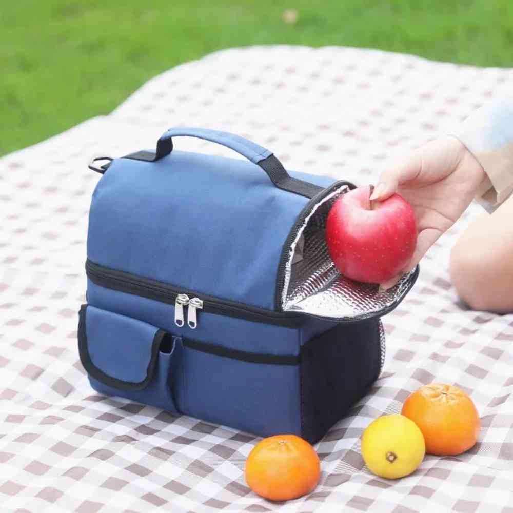  Bolsa Térmica Grande para Marmita com Duplo Compartimento 10L Urbanno azul, aberta em toalha de piquenique, com mão colocando uma maçã no compartimento superior, cercada por frutas cítricas, ideal para lazer ao ar livre urbanno