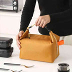 Bolsa Térmica Minimalista Urbanno laranja, sendo segurada por mãos em bancada de cozinha com talheres e pote térmico, demonstrando uso