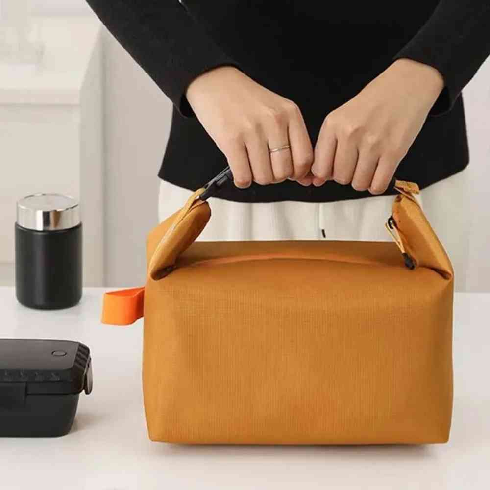 Bolsa Térmica Minimalista Urbanno laranja, sendo segurada por mãos fechada em bancada, com pote e marmita ao lado