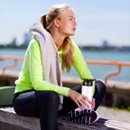 Garrafa de Academia e Hidratação em Alumínio 600ml Urbanno branca, sendo segurada por uma mulher sentada em banco ao ar livre com vista para o mar