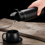 Garrafa de Academia e Hidratação em Alumínio 600ml Urbanno preta, água sendo derramada da garrafa em uma xícara preta, em fundo de madeira