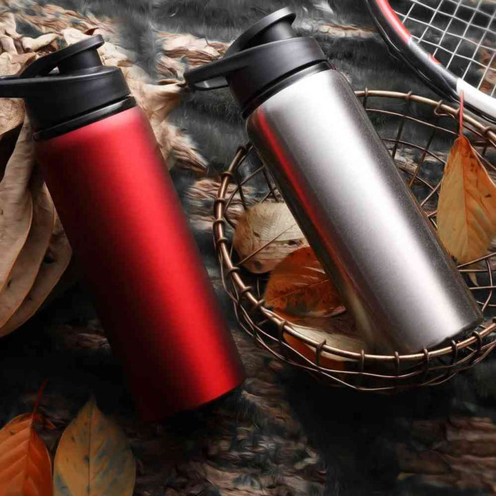 Duas Garrafas de Academia e Hidratação em Alumínio 600ml Urbanno, uma vermelha e outra prata, em cesta com folhas secas e raquete de tênis ao fundo