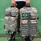 Duas Mochilas Exército Militar Tática 50L ACU (Digital Camo) Urbanno sendo carregadas pelas alças superiores por uma pessoa, com patches das bandeiras dos EUA e do Brasil, demonstrando seu tamanho e facilidade de transporte.