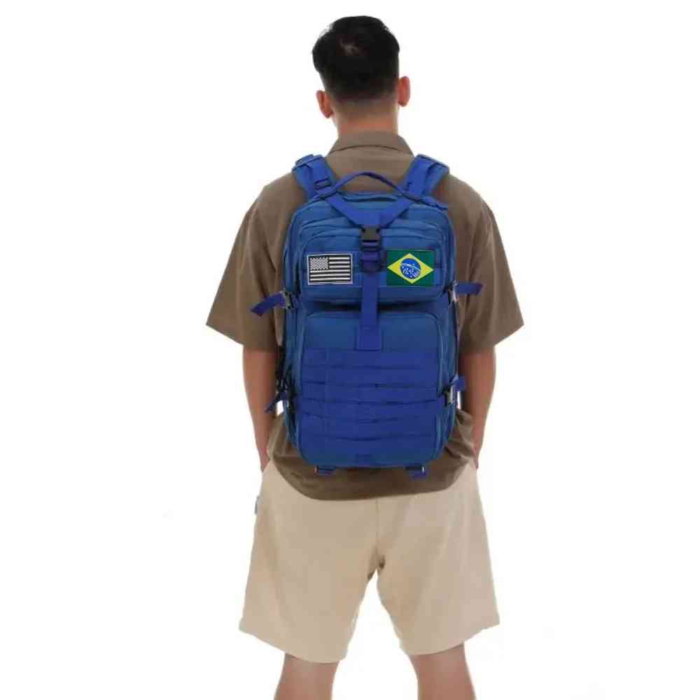 Mochila Exército Militar Tática 50L Azul Urbanno, sendo usada nas costas por um homem de camisa marrom e bermuda, com patches das bandeiras do Brasil e dos EUA, demonstrando o ajuste e o design.