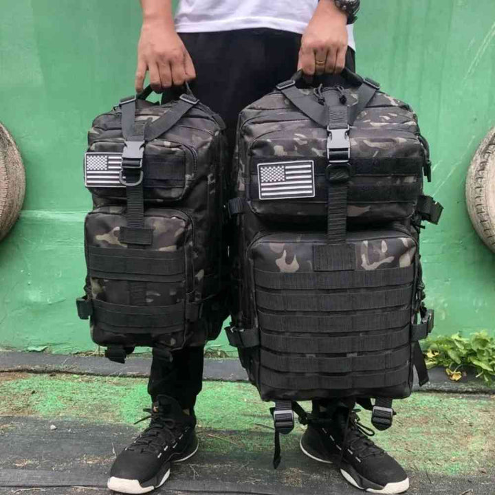 Duas Mochilas Exército Militar Tática 50L Camuflagem Preta Urbanno sendo carregadas pelas alças superiores por uma pessoa, com patches da bandeira dos EUA, demonstrando seu tamanho e facilidade de transporte.
