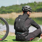 Mochila de Hidratação 10L WEST BIKING com Reservatório 2L Ultraleve Preta Urbanno, sendo usada por um ciclista de costas, sentado na grama com sua bicicleta, demonstrando o ajuste ergonômico e o conforto durante atividades como o ciclismo.
