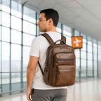 Homem vestindo camiseta branca usando a Mochila Notebook 15.6" em Couro PU Minimalista USB Marrom Tabaco Urbanno nas costas, em um ambiente moderno com grandes janelas, demonstrando seu estilo casual e funcional.