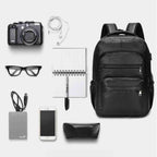 Mochila Notebook 15.6" em Couro PU Minimalista USB Preta Urbanno, com câmera, fones de ouvido, óculos, notebook, celular e outros itens dispostos ao lado, demonstrando sua capacidade de organização.