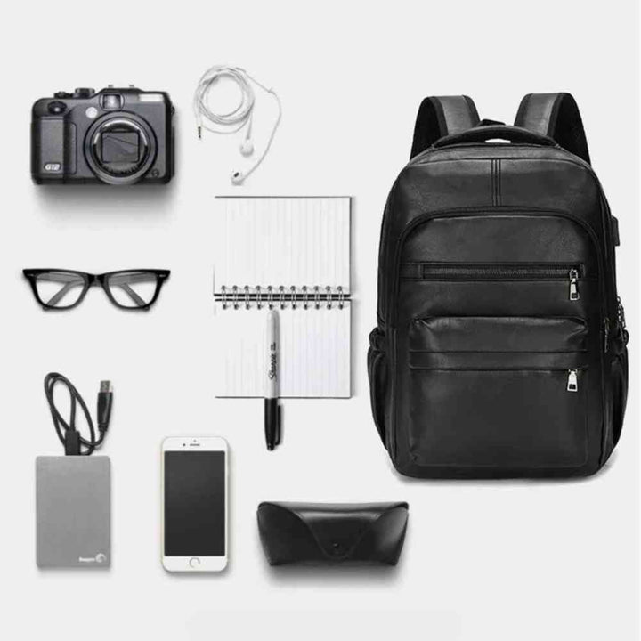 Mochila Notebook 15.6" em Couro PU Minimalista USB Preta Urbanno, com câmera, fones de ouvido, óculos, notebook, celular e outros itens dispostos ao lado, demonstrando sua capacidade de organização.
