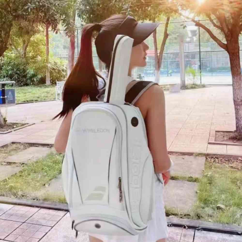 Mulher de costas usando a Mochila Raqueteira Babolat Branca e Dourada Urbanno, com uma raquete inserida e visual de quadra de tênis ao fundo, demonstrando uso para esporte urbanno