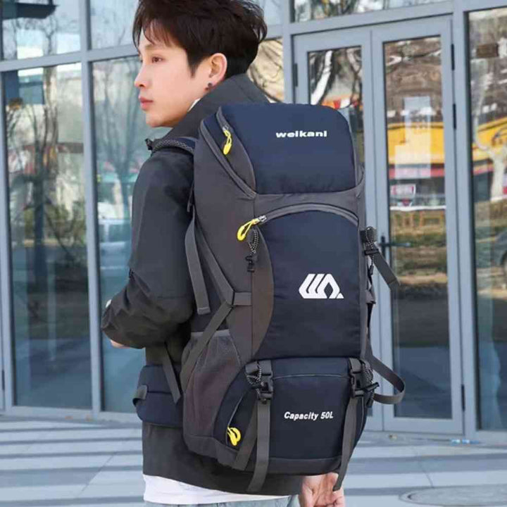 Homem jovem usando a Mochila de Viagem e Camping 50L Aventura Impermeável Urbanno azul nas costas, em ambiente urbano com prédios de vidro ao fundo, demonstrando o tamanho e como veste para aventuras urbanno