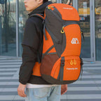 Homem jovem usando a Mochila de Viagem e Camping 50L Aventura Impermeável Urbanno laranja com cinza nas costas, em ambiente urbano com prédios de vidro ao fundo, demonstrando o tamanho e como veste para aventuras urbanno