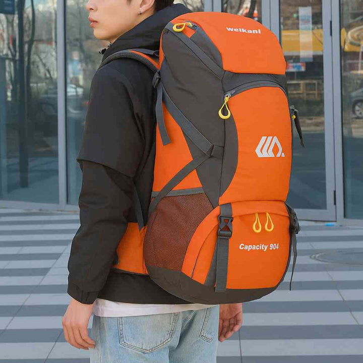 Homem jovem usando a Mochila de Viagem e Camping 50L Aventura Impermeável Urbanno laranja com cinza nas costas, em ambiente urbano com prédios de vidro ao fundo, demonstrando o tamanho e como veste para aventuras urbanno