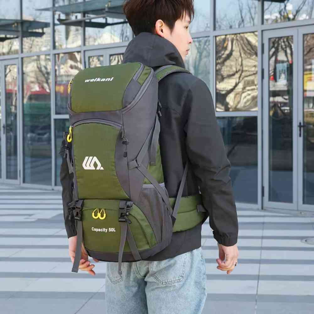 Homem jovem usando a Mochila de Viagem e Camping 50L Aventura Impermeável Urbanno verde oliva nas costas, em ambiente urbano com prédios de vidro ao fundo, demonstrando o tamanho e como veste para aventuras urbanno