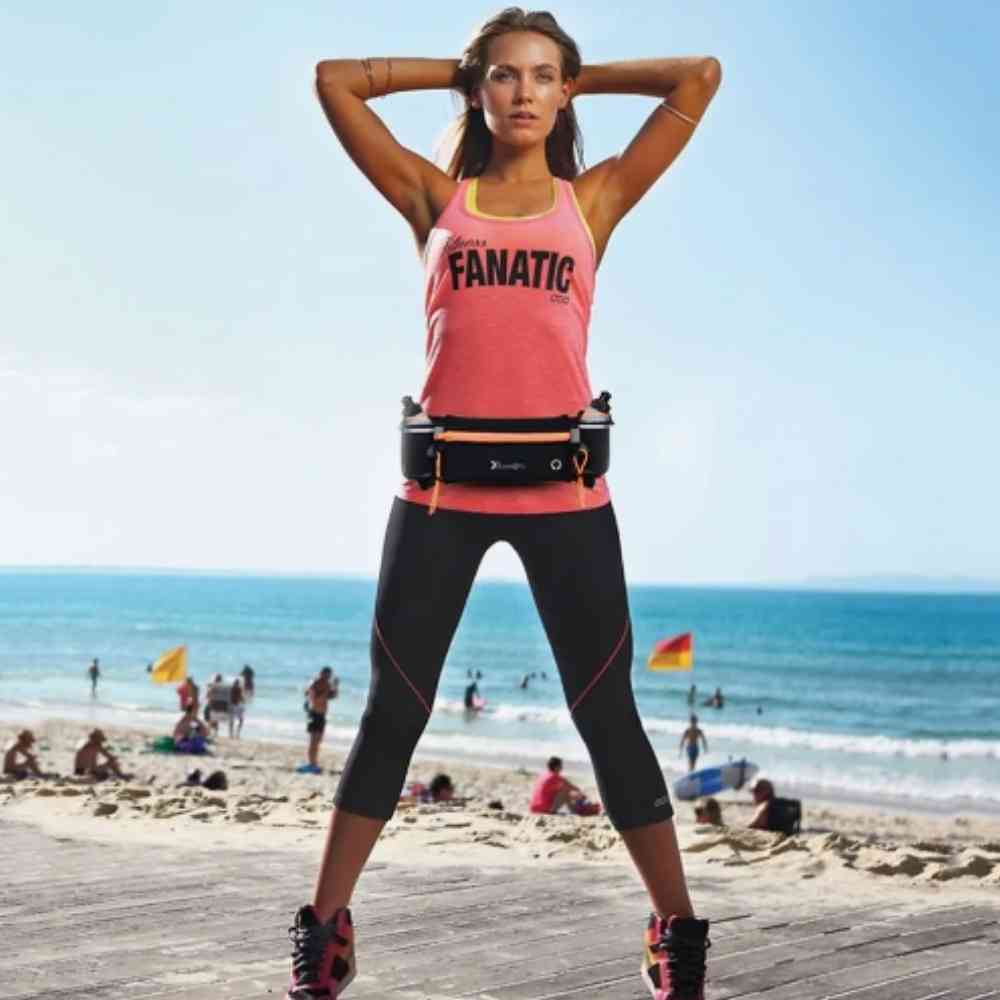 Mulher atlética em roupa de treino preta e regata salmão usando uma pochete de hidratação laranja na cintura, com as mãos atrás da cabeça, em uma passarela de madeira na praia com o mar ao fundo.