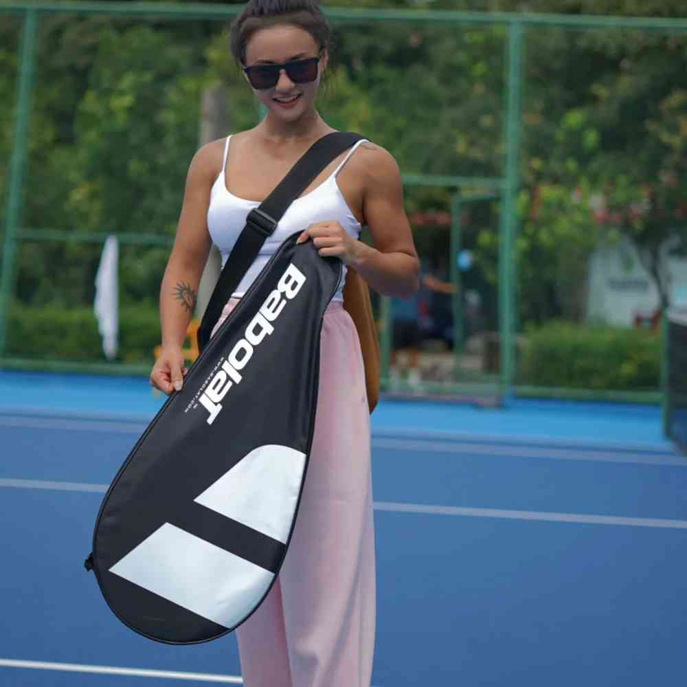 Mulher com óculos escuros usando a Raqueteira Babolat 1 Raquete preta no ombro em uma quadra de tênis, demonstrando o transporte fácil e confortável urbanno