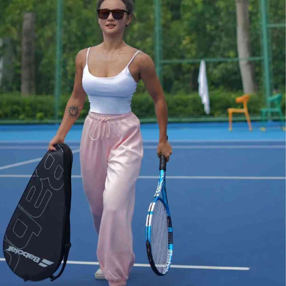 Mulher com óculos escuros segurando a Raqueteira Babolat 1 Raquete preta e uma raquete azul em uma quadra de tênis azul, pronta para jogar, demonstrando uso prático urbanno